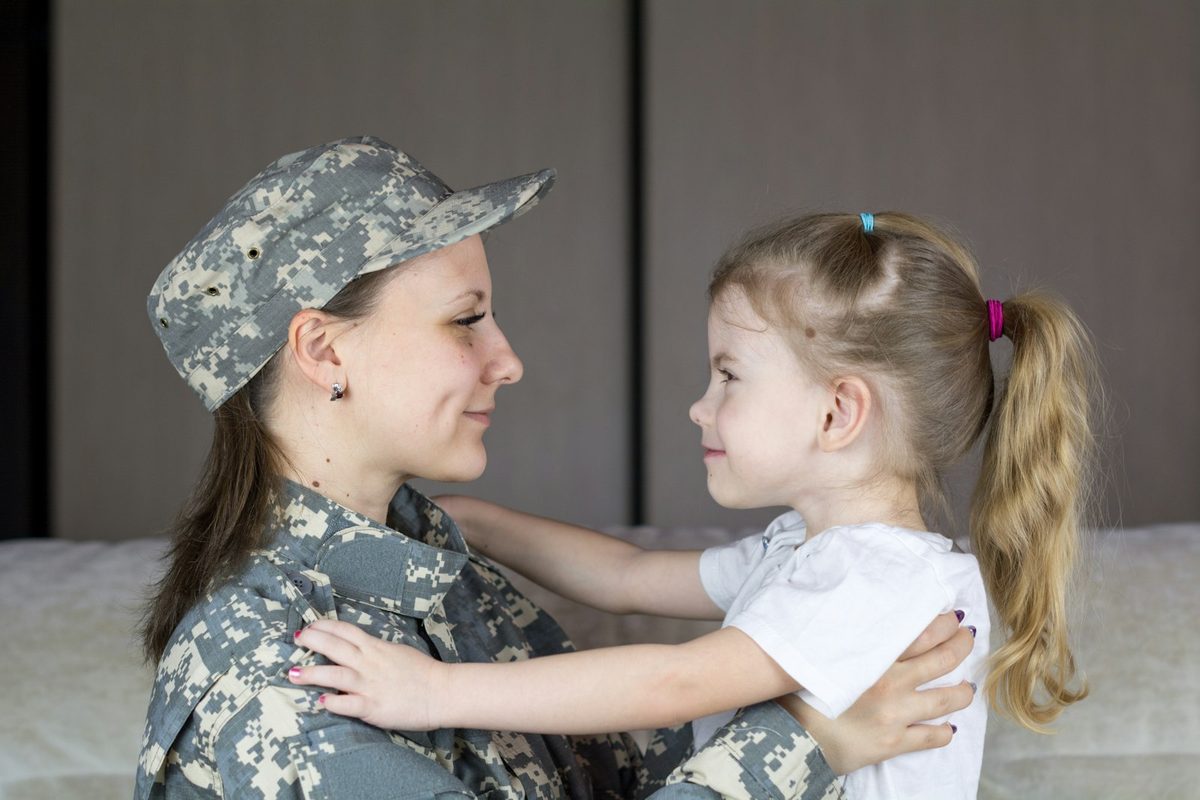Service member in uniform hugging her young daughter at home